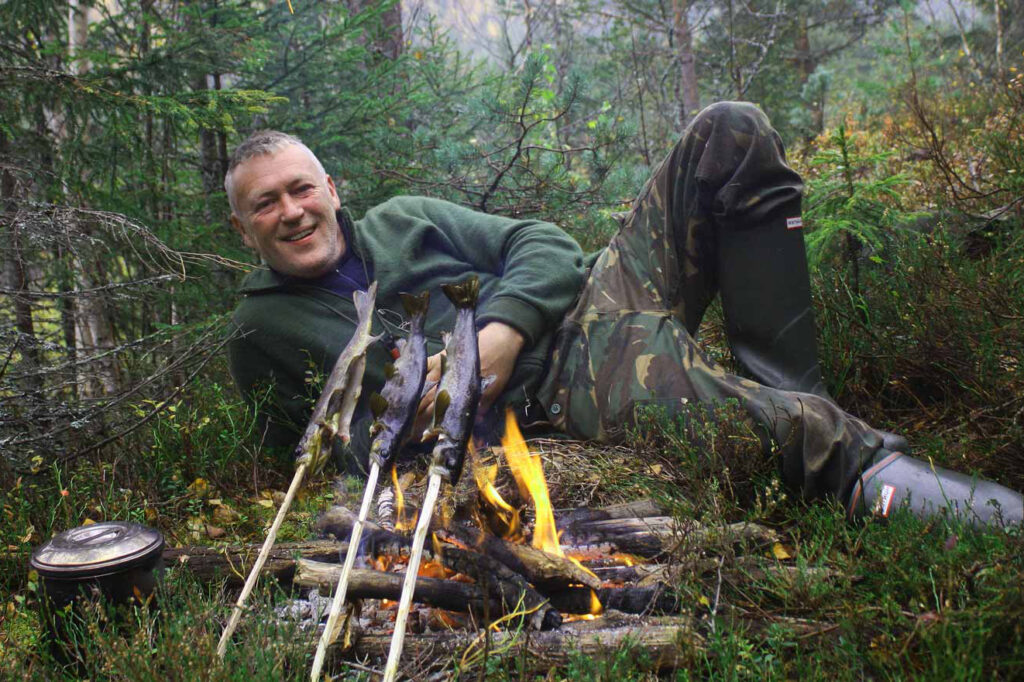 Man lying in front of fire while cooking fish on a stick, bushcraft
