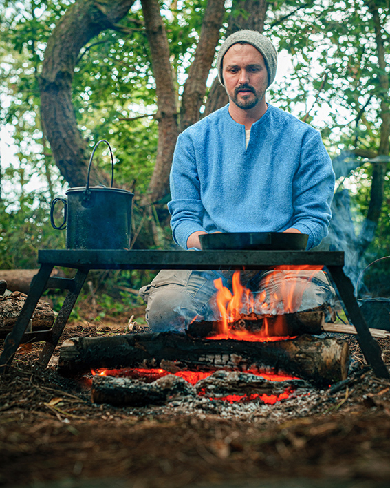 Man with blue wool shirt kneeling in front of campfire with grill over it.