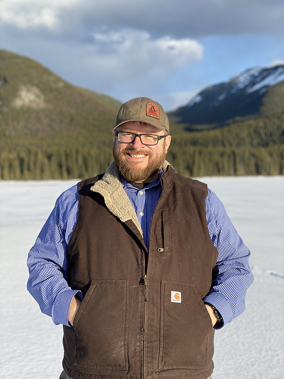 Man with beard standing in front of frozen lake, hills and forest